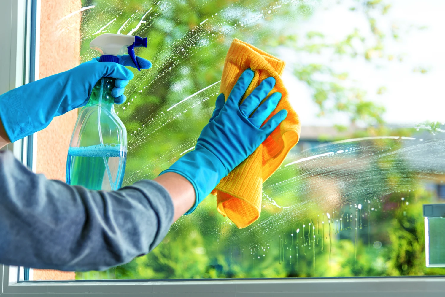 Close-up of a maid wearing blue gloves cleaning a window pane with spray and a yellow cloth