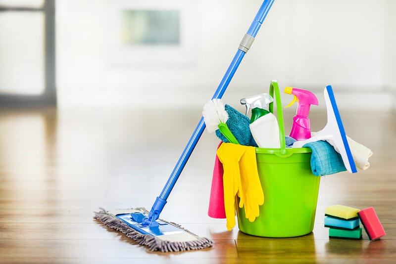 A sweeper and neon green bucket with colorful cleaning supplies set against a blurred out home background